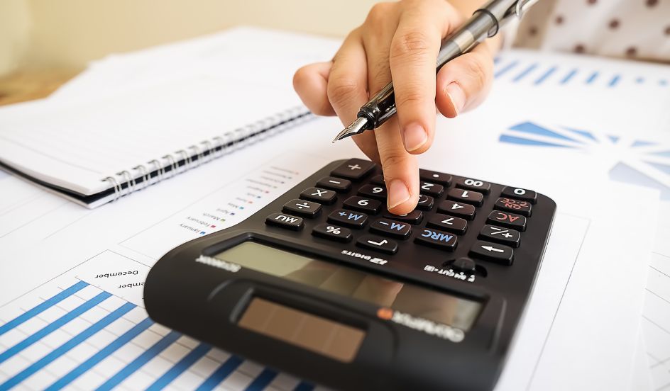 closeup of someone holding a pen as they type into a calculator that rests on a desk covered in paper depicting graphs
